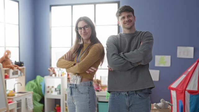 Man and woman teachers stand with arms crossed among toys and a play tent in a bright daycare building, smiling and confident; friendly teamwork.