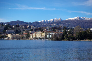 Fototapeta premium Under clear daylight, Lake Lugano in Southern Switzerland, shimmers between mountain slopes, reflecting quiet villages, deep blue water, and the calm rhythm of the alpine landscape.