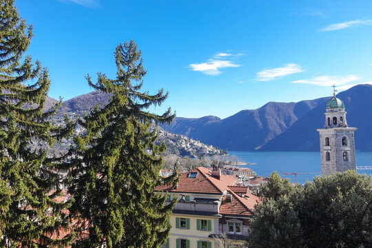 Under clear daylight, Lake Lugano in Southern Switzerland, shimmers between mountain slopes, reflecting quiet villages, deep blue water, and the calm rhythm of the alpine landscape.