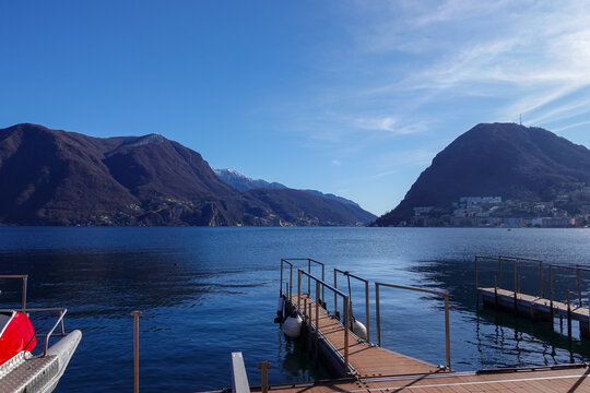 Under clear daylight, Lake Lugano in Southern Switzerland, shimmers between mountain slopes, reflecting quiet villages, deep blue water, and the calm rhythm of the alpine landscape.