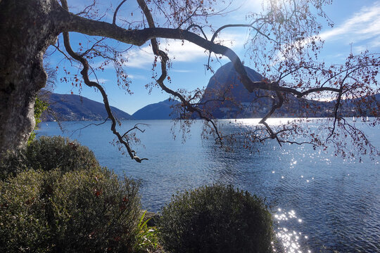 Under clear daylight, Lake Lugano in Southern Switzerland, shimmers between mountain slopes, reflecting quiet villages, deep blue water, and the calm rhythm of the alpine landscape.