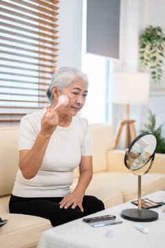 Elderly old asian woman grandma doing make up looking at mirror aside cosmetic while sitting on sofa