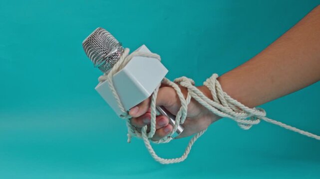 Hand Tightly Holding News Microphone Bound With Thick Rope Against Blue Background For Suppression Of Press Freedom