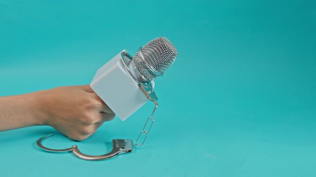 Hand Holding Microphone With Hanging Handcuffs Against Turquoise Background For Arrest And Legal Suppression Of Press Freedom