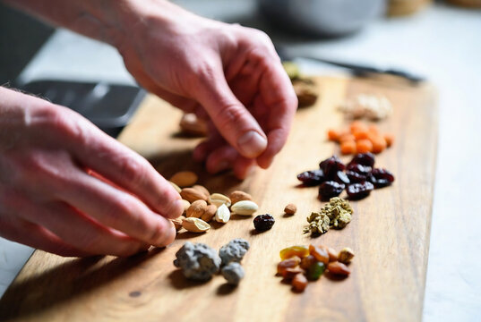Subject: Hands preparing a selection of nuts and dried fruits. Action: Someone is sorting and arranging various nuts and dried fruit on a wooden cutting board. Setting: A kitchen countertop with a lig