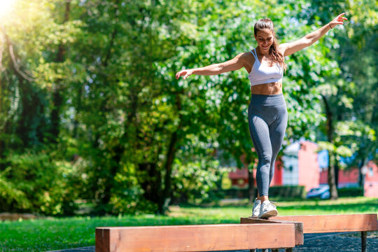 Fit athletic woman balancing with arms extended on wooden beam in park, performing coordination and stability training outdoors.