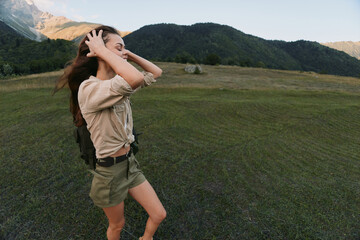 Naklejka premium Woman hiking in a grassy valley, adjusting hair, wearing beige shirt and shorts, backpack ready, surrounded by mountains and wide open sky, vibrant outdoor lifestyle moment