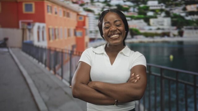 Woman with crossed arms smiling broadly beside a waterfront railing on a street by an orange building and marina; joy confidence.