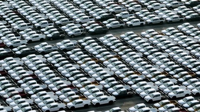 Aerial drone view of thousands of cars parked in organized rows at a large automotive facility, representing global logistics, industrial efficiency, and modern transportation infrastructure. 4k.
