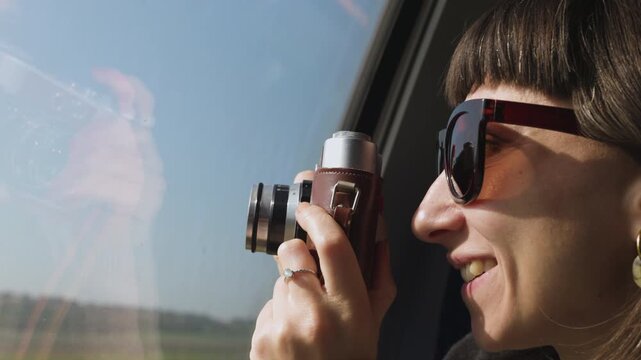 A woman photographs the countryside from a train window. A close-up of a smiling passenger wearing sunglasses holding a camera. Train travel. The passenger enjoys the journey.