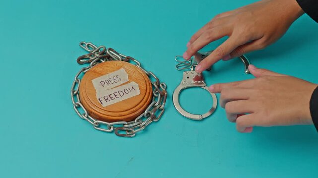 Hand Unlocks Pair Of Metal Handcuffs Positioned Next To Chained Judge'S Sound Block Labeled "Press Freedom" On Blue Background