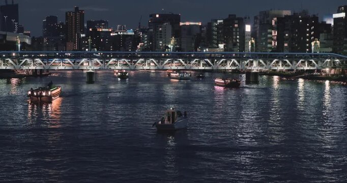 Wide static view, glowing night cityscape over river. Illuminated urban city architecture. Modern building in downtown dark. Cruise boats sailing on sumida river under bridge. Mesmerizing town evening
