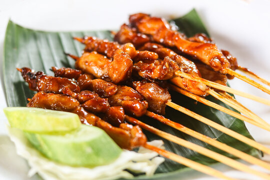 Closeup of marinated grilled chicken satay served on a plate. The chicken is charred and glazed with a savory, slightly sweet sauce. Accompanying the skewers is a side of cucumber slices.
