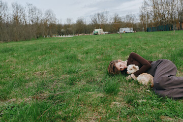 Naklejka premium A woman lies on the grass in a park, cuddling a dog as soft sunlight washes the scene, conveying relaxation, companionship, lifestyle, park, tranquil, candid moments outdoors.