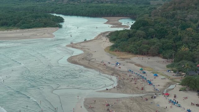 Aerial semi orbit capturing Tamarindo river mouth meeting ocean with beach activity and umbrellas.