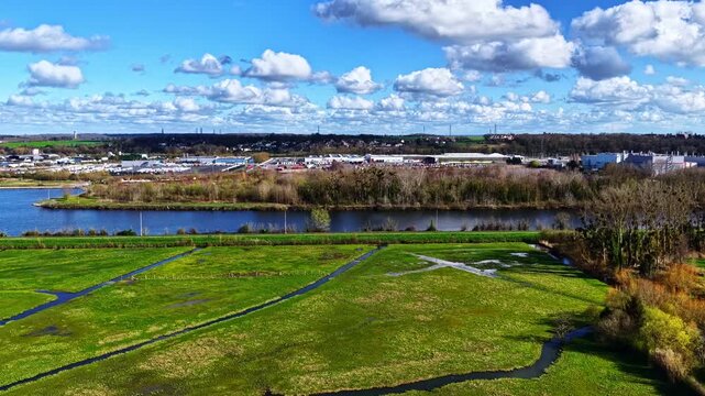 An expansive view of an industrial area beside a river, surrounded by vibrant green fields and under a partly cloudy sky. Useful for environmental studies or urban planning visuals.