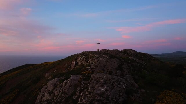 The first light of day casts a gentle glow over a hilltop cross, with pink clouds drifting above the sea. Suitable for spiritual themes or travel content.