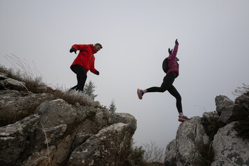 Fototapeta premium Two Hikers Jumping Across Rocky Gorge on Foggy Mountain Trail During Cold Outdoor Adventure