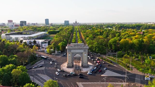 Drone lowering shot tilting up toward Arcul de Triumf, revealing Kiseleff Boulevard and surrounding park during daylight.
