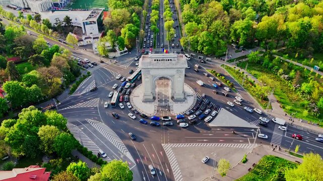 Drone lift and tilt-down shot over Arcul de Triumf roundabout, revealing traffic movement and surrounding greenery of the park.
