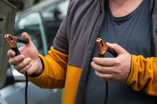 a man's hands holding two black jumper cable clamps, preparing to jump-start a vehicle with a dead battery.