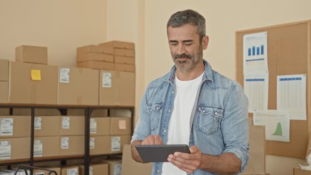 Man tapping tablet while checking packages and boxes in a busy warehouse building; small business focus.