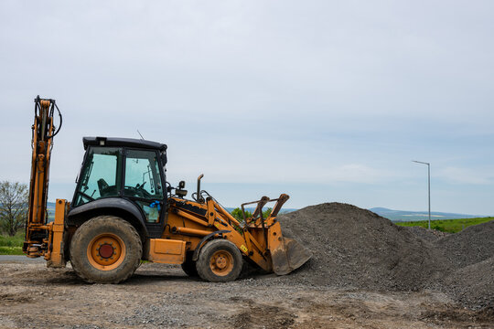 A yellow backhoe loader uses its front bucket to push a large pile of gravel on a dirt lot under a cloudy sky.