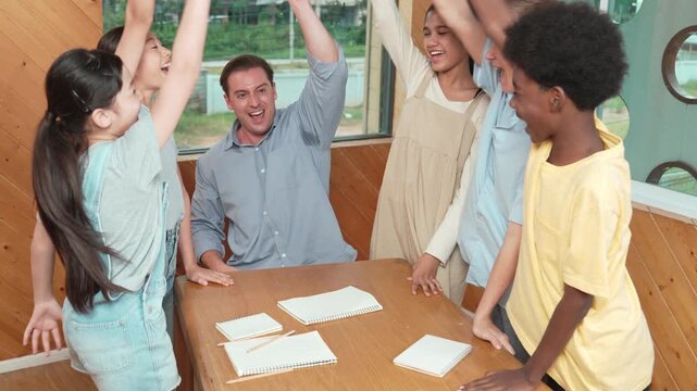 Paper scatter on table while caucasian teacher and diverse children putting hand together to celebrate successful project. Represent love, corporate, team working, togetherness and unity. Edification