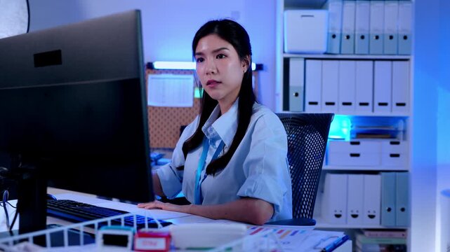 Businesswoman in office working on computer at night overtime, stressed employee at desk with monitor and keyboard, focused night shift workplace scene with paperwork, filing cabinet, modern workspace