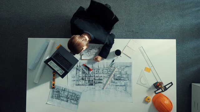 Aerial view of architect drawing while working at laptop. Top down view of caucasian engineer designing blueprint and drafting house model at table with equipment and safety helmet. Alimentation.