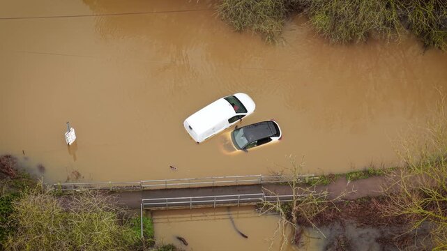 Flooded cars sinking in muddy water during heavy rain season in UK. Severe flooding covering rural road and trapping vehicles near small bridge. Extreme weather causing transport disruption and