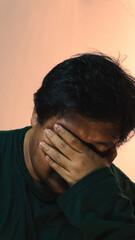 Fototapeta premium A distressed adult male wearing a dark green shirt bows his head and covers his eyes with one hand to show feelings of sadness or deep exhaustion against a warm peach background. Deep