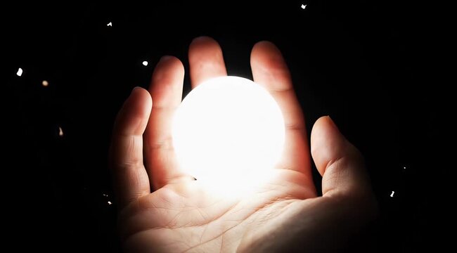 Close up of human hands holding a bright glowing orb of light against a dark background with floating particles and soft warm lighting creating a magical ethereal atmosphere