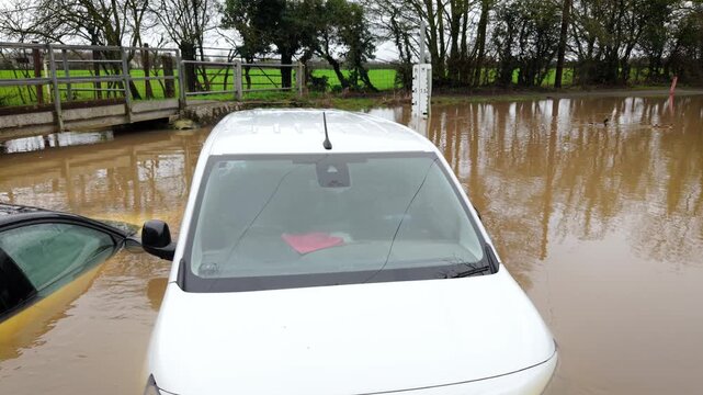 Climate disaster flooding countryside road and submerging parked vehicles. Murky water surrounding transport infrastructure and causing severe property damage. Extreme rainfall intensifying and