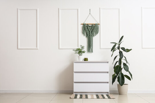 Interior of living room with houseplants, commode and hanging tapestry on white wall
