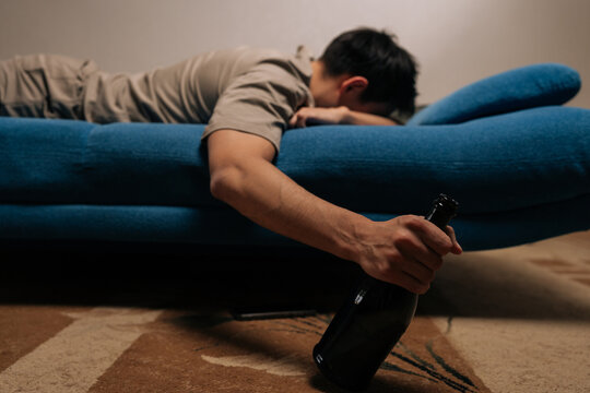 Drunk man suffering from alcohol dependence, lying on blue sofa, holding bottle of alcoholic drink, depicting unconsciousness and devastating impact of substance misuse and long-term abuse.