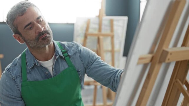 Man in green apron leans and holds a paintbrush to canvas on an easel in an art studio, then rests hands behind head; creative contemplation.