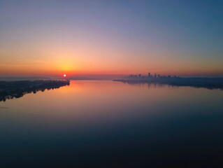 Obraz premium Sunrise over a tranquil river with a distant city skyline emerging from mist