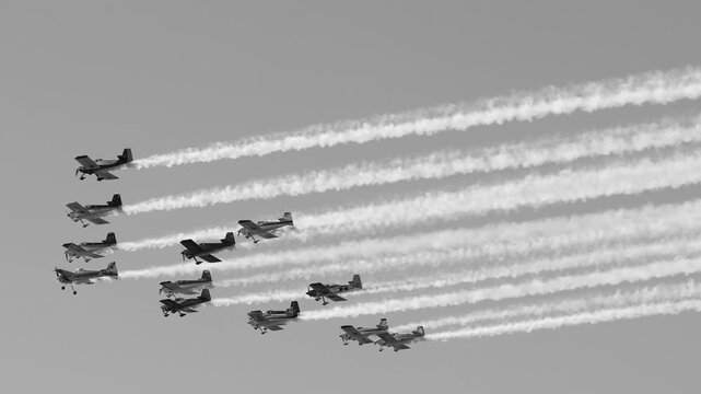 Group of formation planes flying through sky in grayscale with long smoke trails following each aircraft.