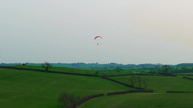 Paraglider gliding above rolling green countryside fields hedgerow farmland under overcast sky. Extreme paramotoring adventure flying high over rural landscape outdoor sport adrenaline freedom. Aerial