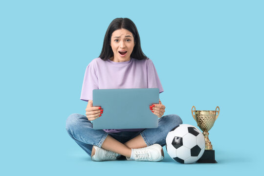 Shocked young woman with laptop and soccer ball sitting on blue background. Sports bet concept