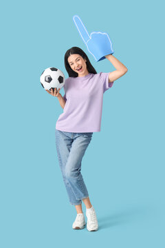 Happy young woman with soccer ball and foam finger on blue background. Sports bet concept