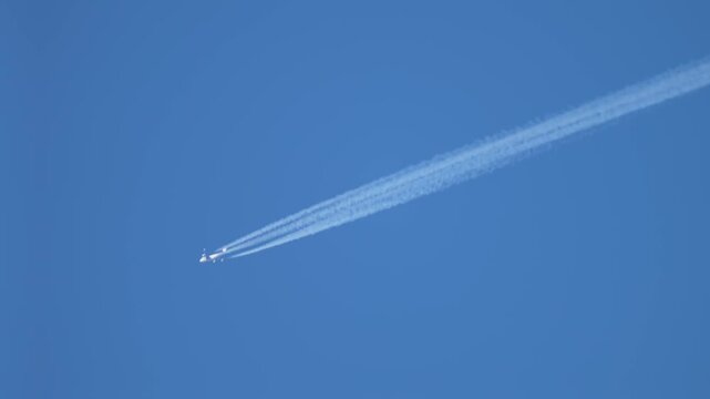 Wide body jet creating long contrail lines across clear blue sky, high altitude aviation traffic and chemtrails concept. Four engine airplane exhaust plume linking air pollution, climate change and