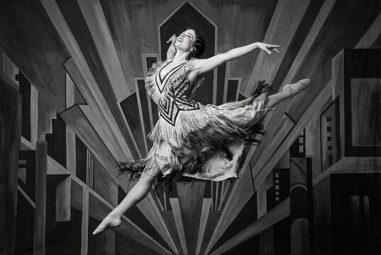 Energetic female ballet dancer mid-air in a flapper fringed dress performing against a painted Art Deco geometric city background in black and white.