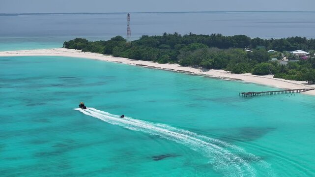 Aerial view of a motorboat towing an inflatable tube across turquoise waters along the white sandy beach and lush tropical vegetation of Vashafaru, Haa Alif Atoll, Maldives.
