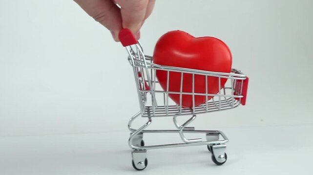 Hand pushes shopping cart with red heart inside. White background highlights the gesture of love. Cart moves slowly, symbolizing care in shopping. Red heart represents emotional value over price