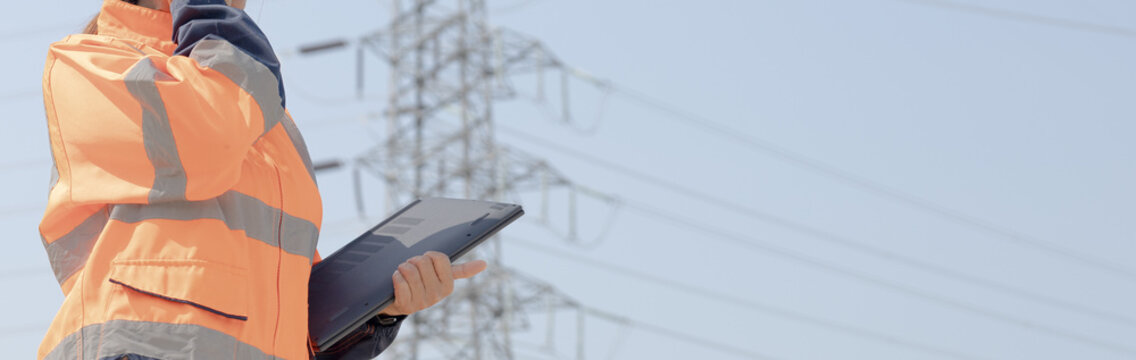 A worker stands outside in bright clothing and looks up at the power lines. They hold a tablet and check details related to the electrical infrastructure