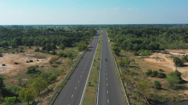 Motorcyclist travels alone on divided highway cutting through rural landscape in Tamil Nadu. Aerial view captures the road flanked by green vegetation and scattered trees under clear sky.