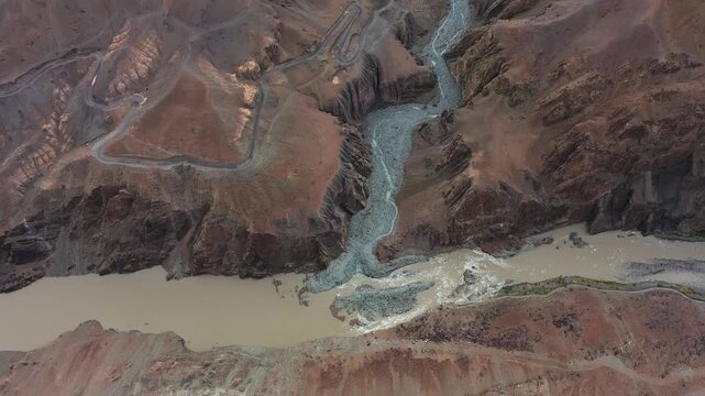 Ladakh's Indus River gorge cuts through barren mountain terrain, revealing sediment patterns and winding roads. Smooth drone movement looks down at glacial confluence point from high altitude.