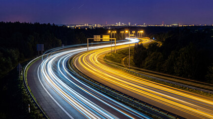 A long exposure photograph of car light trails on a winding highway at night, with distant city lights and streetlamps. © Jacqueline